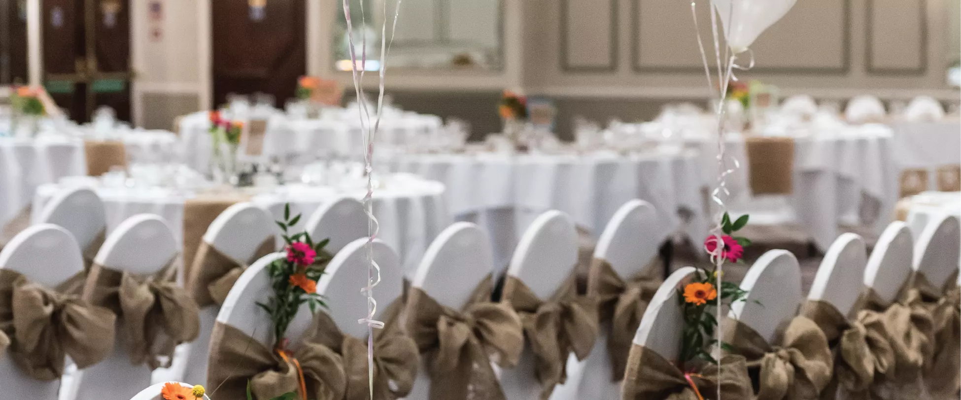 A large room set up for a wedding with chairs covered in white with burlap ribbons, flowers and balloons.