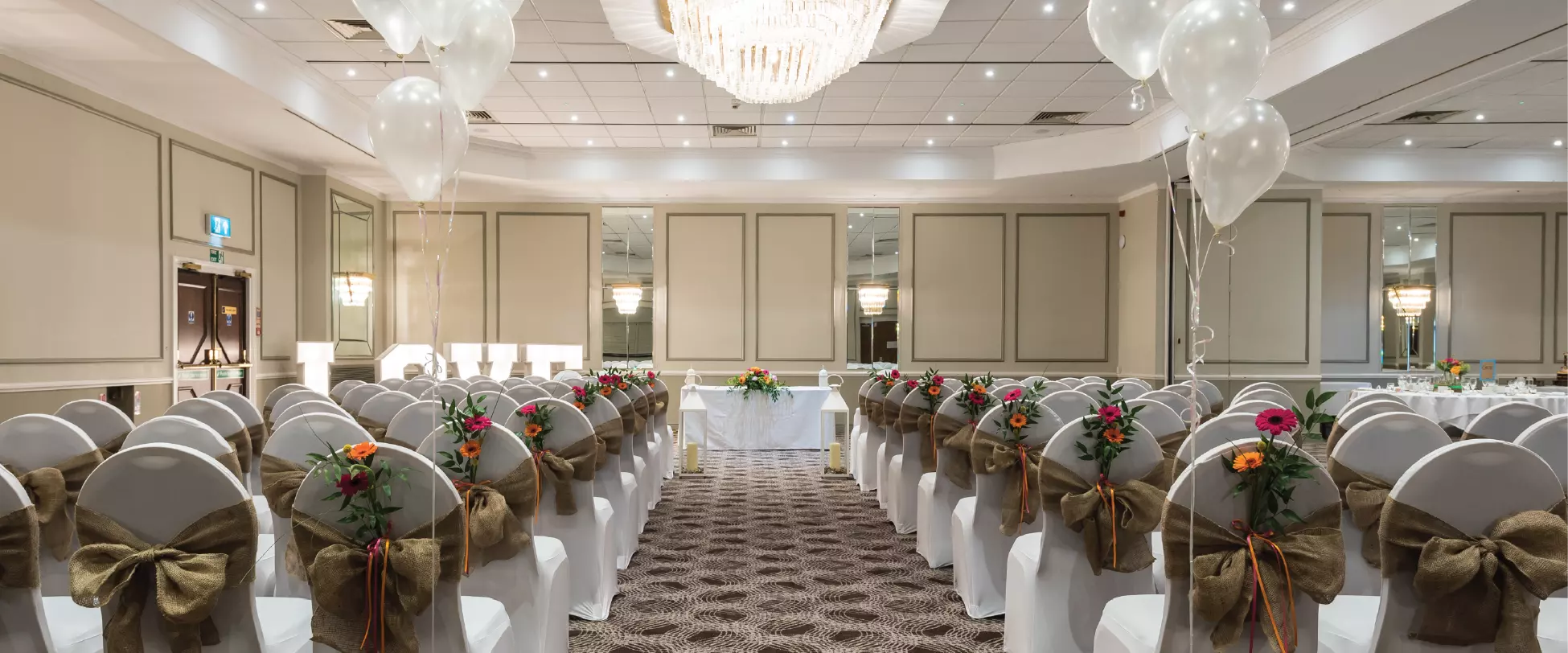 A large room set up for a wedding with chairs covered in white covers, burlap bows, and flowers.